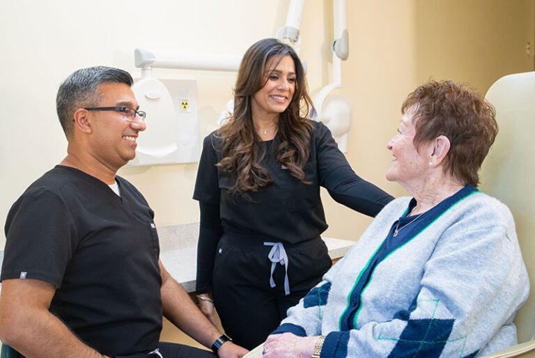 Dental staff smile at an elderly patient seated in a clinic chair, discussing care.