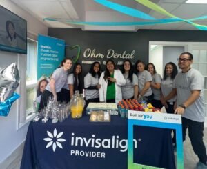 Group of dental staff posing behind an Invisalign promotional table in a dental office with blue tablecloth and banners under hanging streamers.