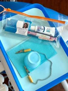 Blue dental tray with a clear zip pouch holding toothpaste and dental care products, plus a blue cap and small clamps on a blue mat (at-home dental kit).